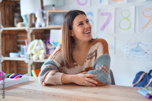 Young beautiful teacher woman wearing sweater and glasses sitting on desk at kindergarten looking away to side with smile on face, natural expression. Laughing confident.