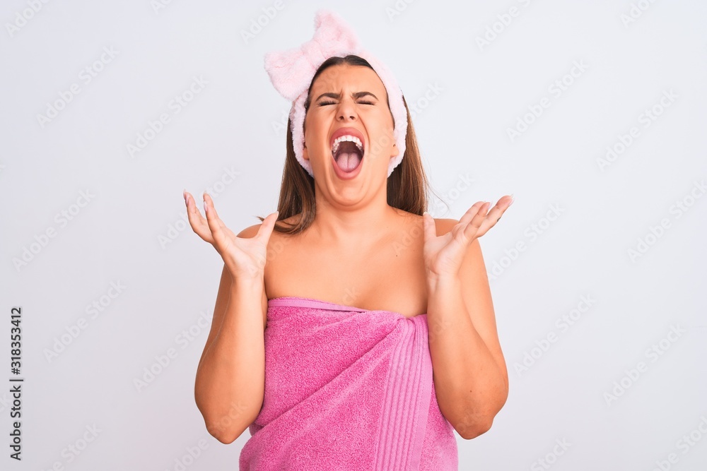 Beautiful young woman wearing pink shower towel and beauty headband over white background crazy and mad shouting and yelling with aggressive expression and arms raised. Frustration concept.