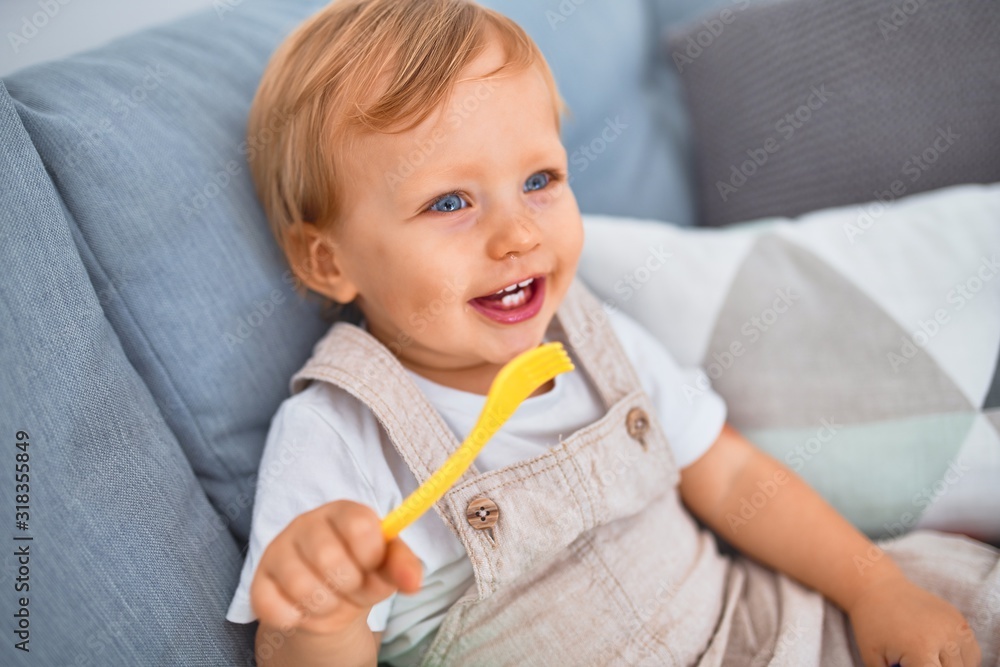 Adorable blonde toddler smiling happy sitting on the sofa playing with plastic meals toys at home - obrazy, fototapety, plakaty 