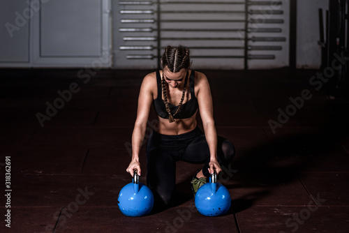 Young sweaty strong muscular fit girl with big muscles sitting on the floor and holding two heavy kattlebell weights with her hands resting after hardcore crossfit workout training in the gym