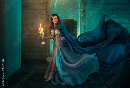 Luxury lady Queen medieval royal dress run escapes from Gothic night castle. Blue silk dress, cloak train plume waving motion. Holds in hands old candlestick burning candles. Background old retro room