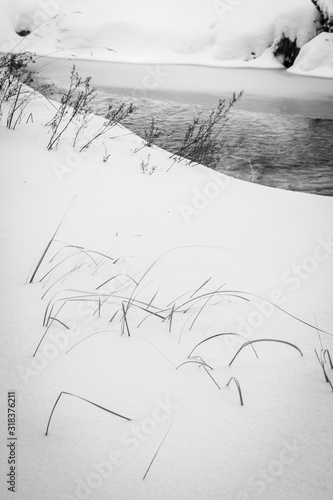 Vertical black-and-white fine art print of grass and shrubs in snow with river frozen over background