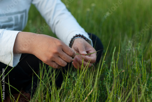 Hands of a woman holding a grass straw in a meadow wearing black trousers and a white shirt.