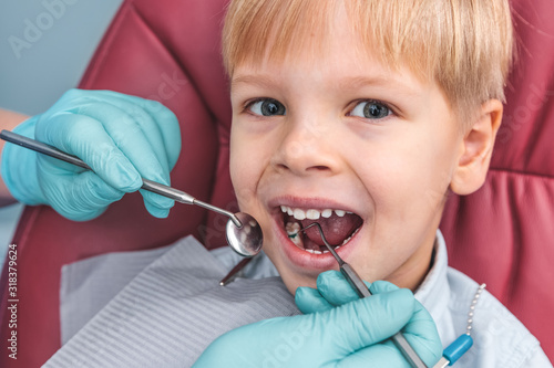 Portrait of happy young boy sitting in dental chair, dentist holding in hands in blue gloves mouth mirror and periodontal explorer scaler, healthy teeth concept