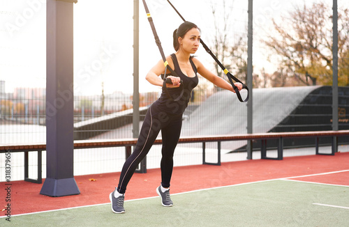 Girl athlete functional training on sportground. Mixed race young adult woman do workout with suspension system. Healthy lifestyle. Stretching outdoors playground. © Andrii