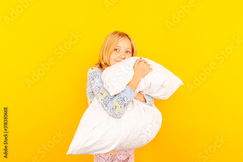 Adorable little girl looking at the camera and hugging a pillow background of a yellow wall.