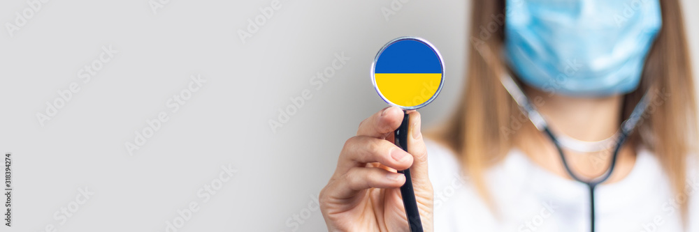 Young woman in a medical mask holds a stethoscope with the flag of Ukraine on a light background. Concept of medicine, virus, epidemic, vaccination. Banner - obrazy, fototapety, plakaty 