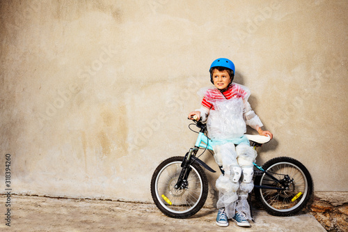 Boy with bicycle ball wear overprotecting super safe bubble wrap cover and helmet stand near the grey wall on the street photo