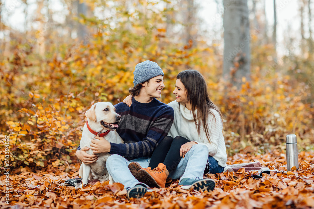 Fototapeta premium Young couple having picnic with drink and their golden labrador in the park, sitting on blanket.