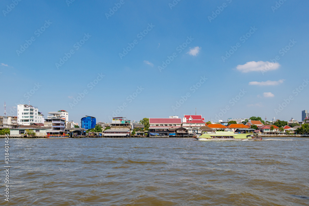 Outdoor sunny view of Express boat on Chao Phraya River and cityscape along waterside against blue sky.