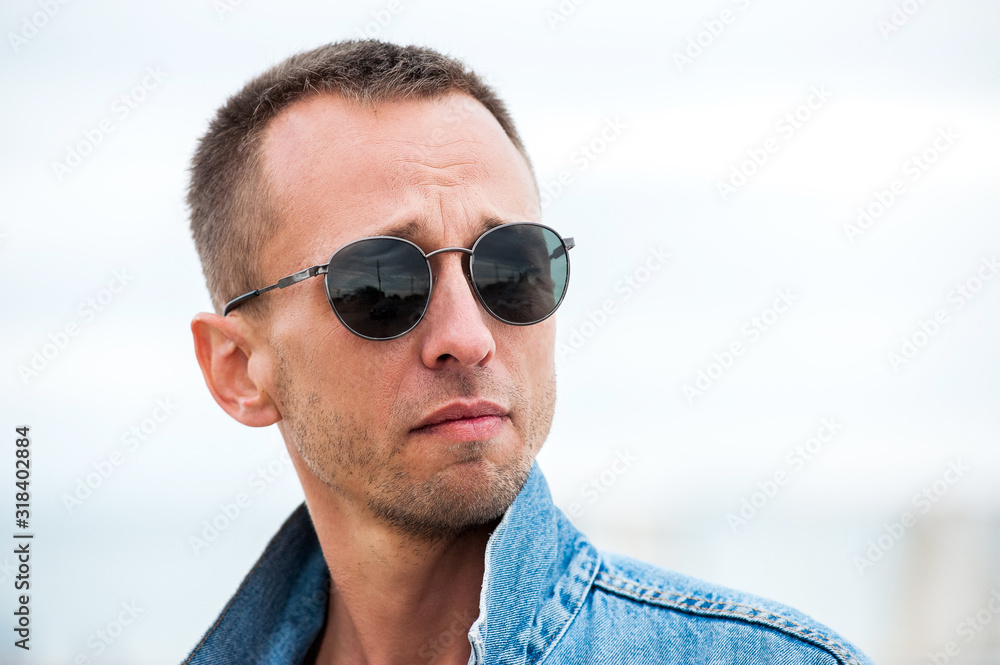 young man in sunglasses and a denim jacket looks away with a serious expression on his face young man in sunglasses and a denim jacket looks away with a serious expression on his face