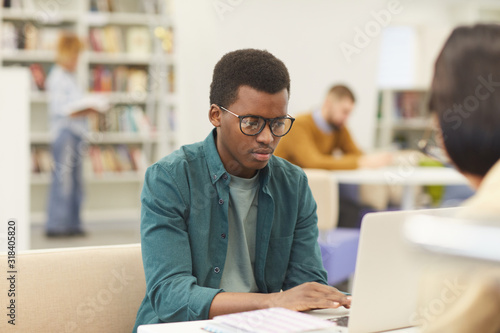 Portrait of African teenage boy studying in college library, using laptop with other students in background, copy space