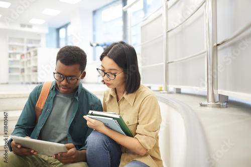 Portrait of two international students reading books together while working on project in modern college library, copy space