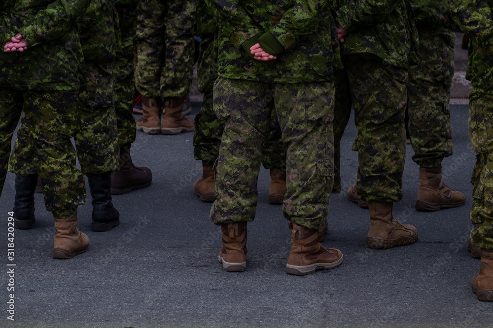 A group of military officers or personnel dressed in green fatigues, camouflage clothing and wearing brown boots. The men have their hands behind their backs standing at ease on black pavement. A group of military officers or personnel dressed in green fatigues, camouflage clothing and wearing brown boots. The men have their hands behind their backs standing at ease on black pavement.