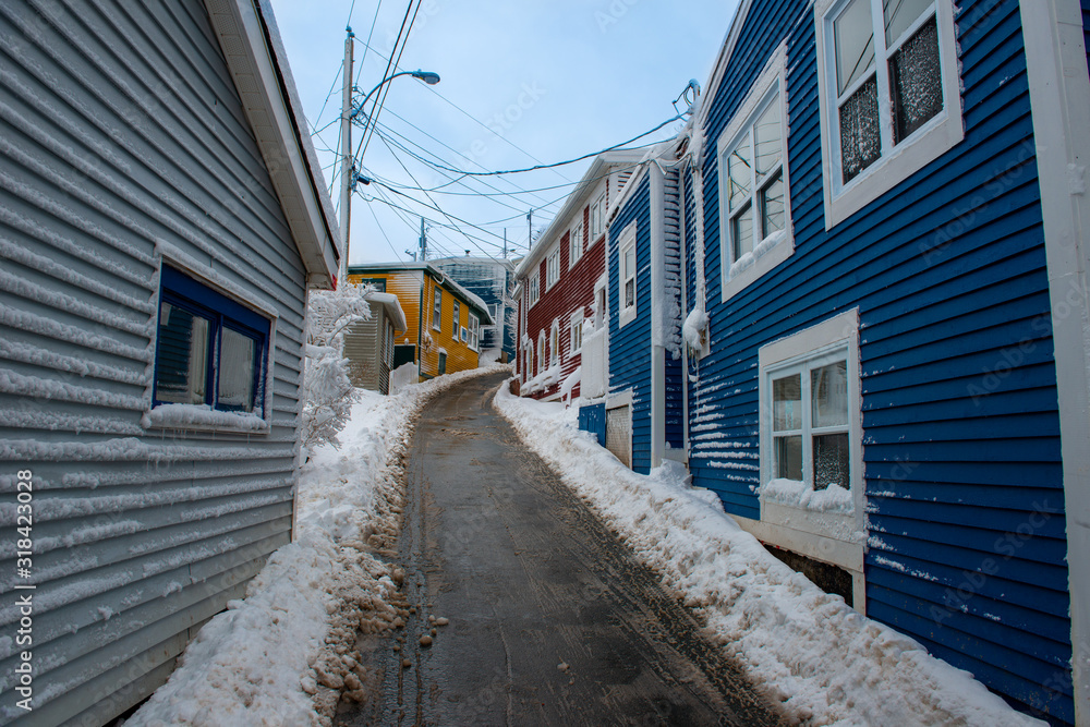 Colorful buildings on a narrow street with snow on both sides of the road. The buildings are blue, red, orange and green made of wood clapboard with windows. There are light poles and wires in the sky