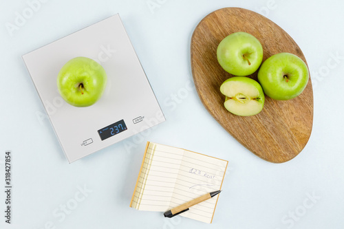 Fresh green apple on gray digital kitchen scales. Near notepad with number of calories, pen and some apples on wooden board. Flat lay. Kitchen equipment. Healthy food concept. Weighing products.