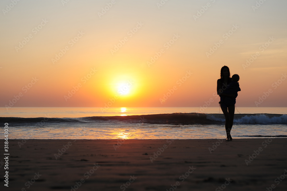Silhouette of woman with a child in her arms on the background of sunset by the sea. Young slender girl carries a baby in the rays of setting sun. Beautiful mother walking along the sandy beach.
