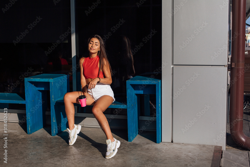 Stylish happy young brunette woman wearing white shorts and sneakers holding pink cup of coffee to go.