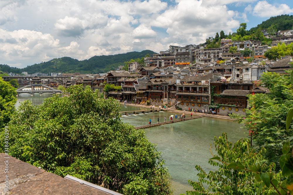 Panoramic view of the old town. Tuojiang River, traditional houses and local chinese people crossing river. Bridge is made of stone blocks.  Fenghuang Town, Hunan province, China