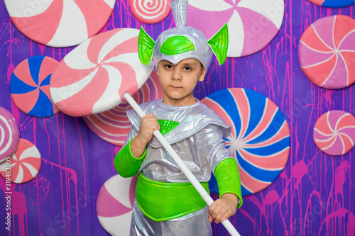 A boy in an alien costume holds a big candy in his hands against the background of a wall of sweets. photo