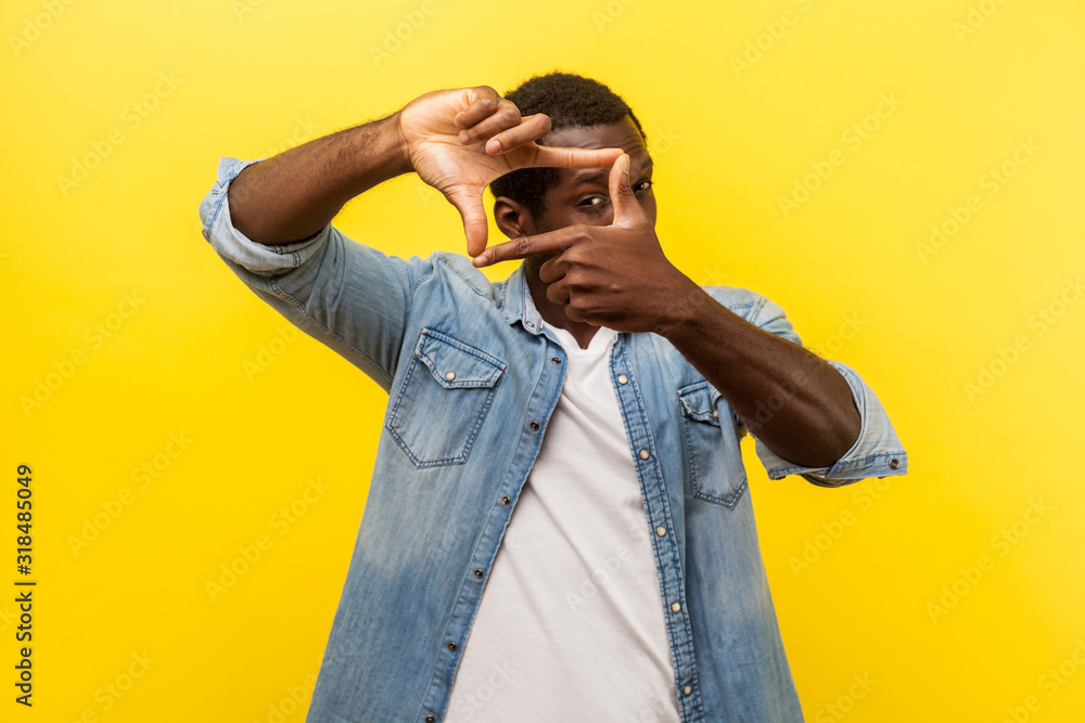 Portrait of focused curious man in denim casual shirt with rolled up sleeves looking through photo frame made of hands, viewing distant with interest. indoor studio shot isolated on yellow background Portrait of focused curious man in denim casual shirt with rolled up sleeves looking through photo frame made of hands, viewing distant with interest. indoor studio shot isolated on yellow background