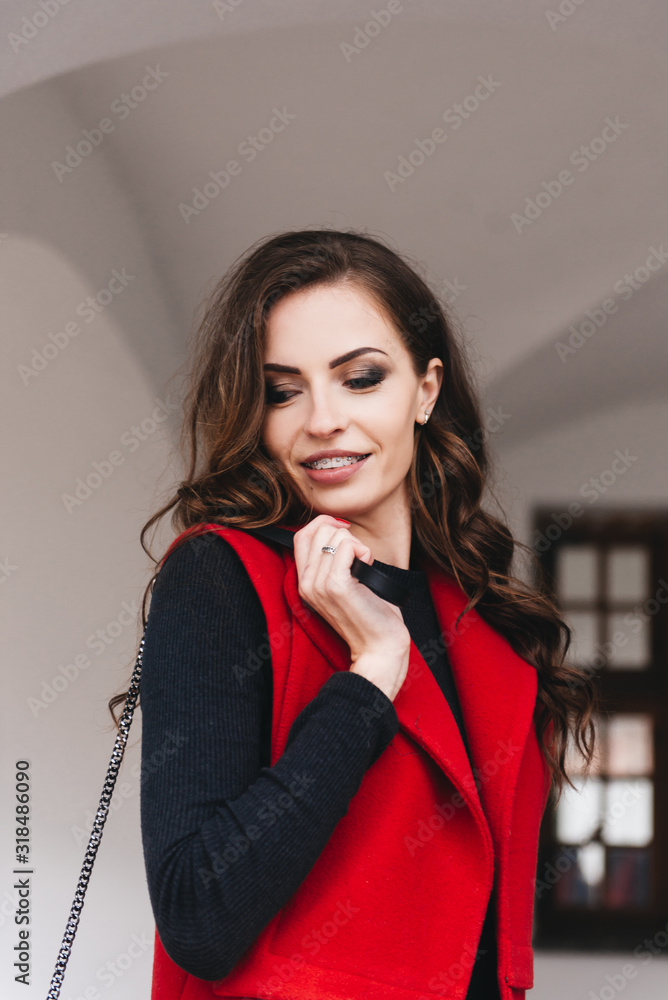 Outdoor portrait of a stylish young girl on the street of an old city in black clothes and a long red vest. A model poses on the street of a European city