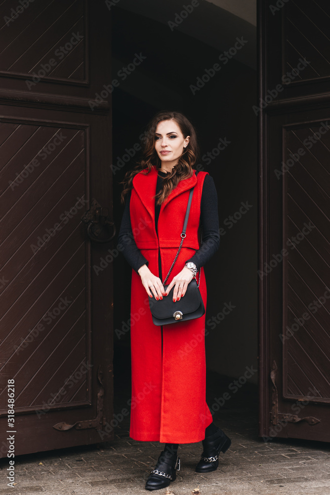 Outdoor portrait of a stylish young girl on the street of an old city in black clothes and a long red vest. A model poses on the street of a European city