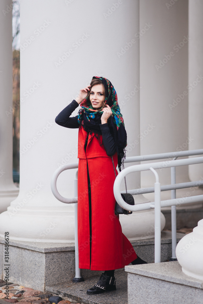 Outdoor portrait of a stylish young girl on the street of an old city in black clothes and a long red vest and a Russian scarf on her head. A model poses on the street of a European city