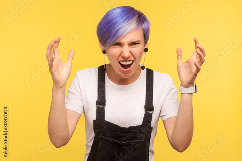 Portrait of aggressive hipster girl with violet dyed short hair in denim overalls quarreling with raised hands, screaming in anger with mad and furious face. isolated on yellow background, studio shot