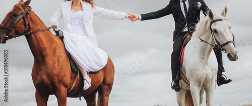 The groom in a black suit and the bride in a white dress are sitting on horses, walking and holding hands. Wedding portrait of the newlyweds with beautiful animals. Photography, concept.