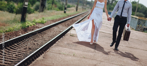 Happy newlyweds are walking on the platform holding hands, waiting for the train and honeymoon. The bride and groom go on vacation to warm countries. Photography, concept.