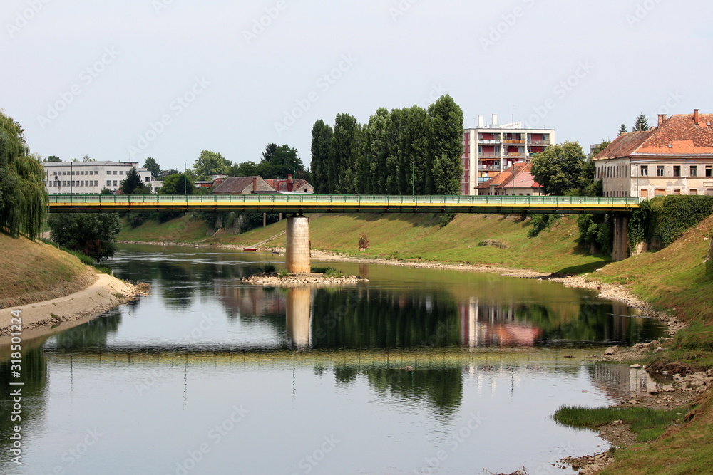 Green and yellow old metal bridge mounted on strong concrete foundation over local river with low summer water levels surrounded with apartment buildings and family houses among tall trees