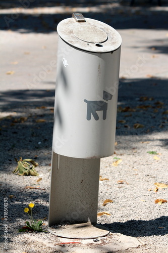Grey metal dog waste bin with closed top mounted on concrete foundation surrounded with small grain gravel in local public park