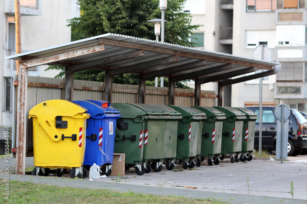 Row of various large plastic trash containers for ecologically sorting garbage for recycling under metal roof tiles protection between old tall apartment buildings Row of various large plastic trash containers for ecologically sorting garbage for recycling under metal roof tiles protection between old tall apartment buildings