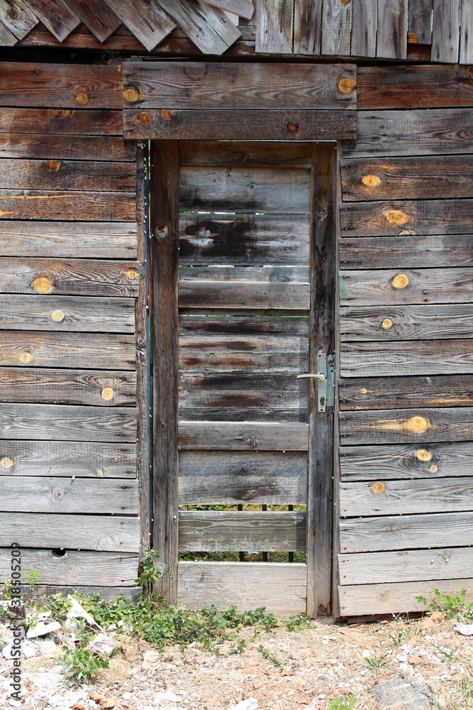 Tall narrow cracked wooden boards doors mounted on abandoned suburban family house backyard entrance with grass and gravel in front Tall narrow cracked wooden boards doors mounted on abandoned suburban family house backyard entrance with grass and gravel in front