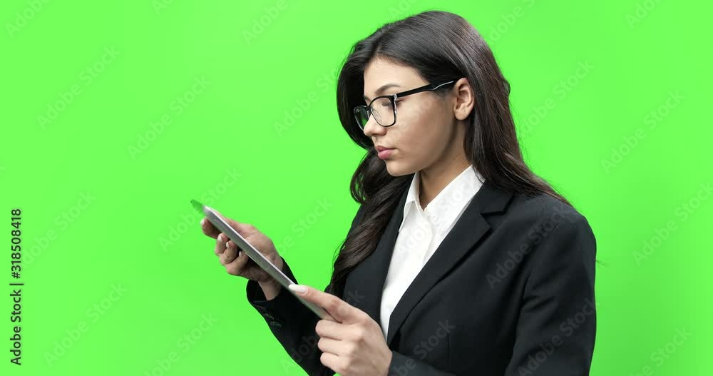 Side view of beautiful serious woman in black suit and eyeglasses looking at digital tablet while standing against green background. Concept of technology, business and finance. Side view of beautiful serious woman in black suit and eyeglasses looking at digital tablet while standing against green background. Concept of technology, business and finance.