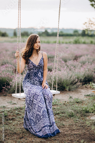 beautiful girl in a long dress sits on a swing in a lavender field at sunset. Soft focus.