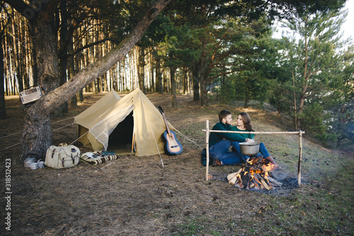 Young loving couple of tourists camping, sitting near a campfire against the tent in forest