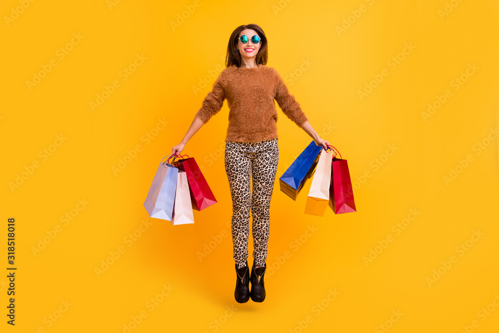 Full body photo of funny overjoyed lady jump high carry packs addicted shopaholic wear sun specs casual fluffy sweater leopard trousers boots isolated yellow color background Full body photo of funny overjoyed lady jump high carry packs addicted shopaholic wear sun specs casual fluffy sweater leopard trousers boots isolated yellow color background