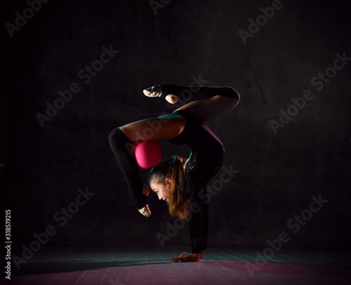 Young girl gymnast in black sport body and uppers standing in bridge pose and holding pink gymnastic ball between leg and neck