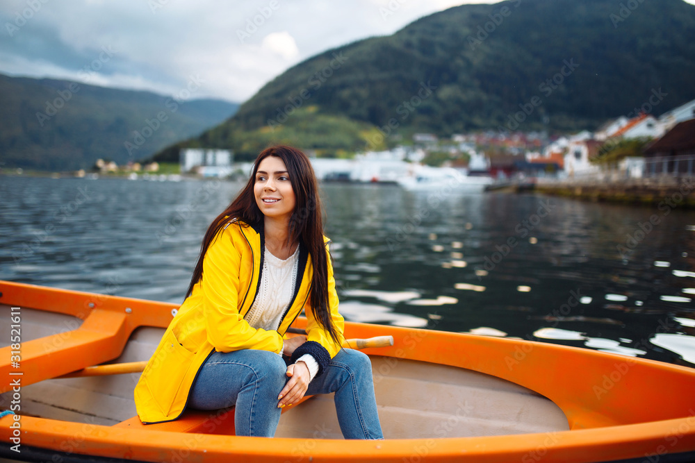 The girl tourist in a yellow jacket  is sitting in a boat against the backdrop of the mountains in the Norway.  Active woman relaxing on the boat by the lake. Travelling, lifestyle, adventure concept.