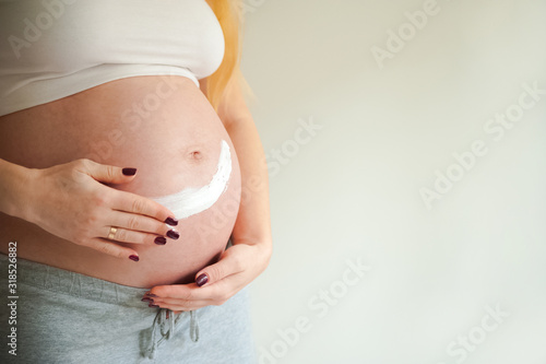 A pregnant woman is applying cream to her stomach for stretch marks. Pregnant tummy, oil for skin elasticity. Pregnant cares for her stomach on a gray background close-up and copy space. © Nelly