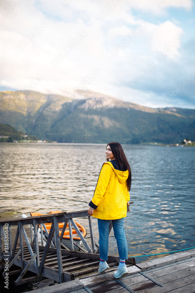 The girl tourist in a yellow jacket posing on the lake in Norway. Active woman relaxing near the lake against the backdrop of the mountains in the Norway. Travelling, lifestyle, adventure, wild nature