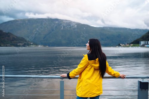 The girl tourist in a yellow jacket posing on the lake in Norway. Active woman relaxing near the lake against the backdrop of the mountains in the Norway. Travelling, lifestyle, adventure, wild nature