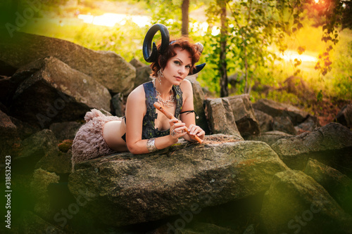 A girl in the image of a faun lies on stones with a wooden flute. Model with horns on the head and with furry pants in the forest photo