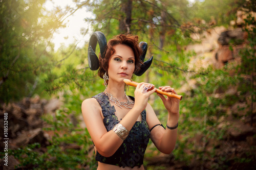 A girl in the image of a faun stands with a wooden flute. Model with horns on his head and with furry pants in the woods among the stones photo