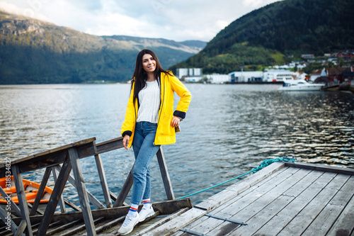 The girl tourist in a yellow jacket posing by the lake in Norway. Active woman relaxing near the boat by the lake against the backdrop of the mountains in the Norway. Travelling, lifestyle, adventure. © maxbelchenko