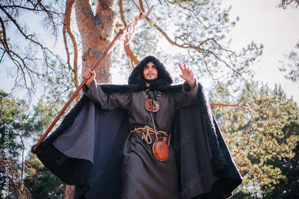 A man with a beard and a mustache in a robe and coat with a staff in his hand. A magician, a shaman conjures in the forest. Fantasy image