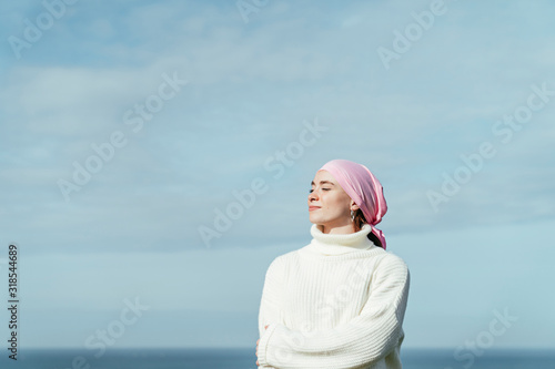 Portrait of young woman with cancer and crossed arms with closed eyes and sky on background