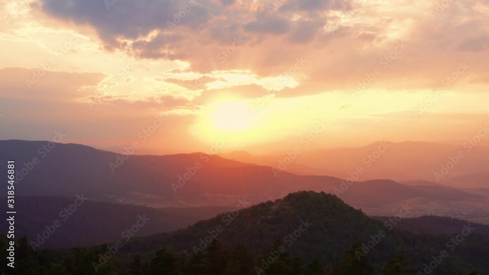 Lens flare sun through tops of fir trees on panorama of mountains landscape at orange sunset and bright disk of sun in evening in summer against blue sky with clouds aerial view. Drone ahead. Nature